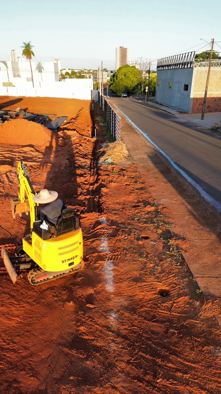 Mini escavadeira abrindo vala ao longo da via, vista aérea